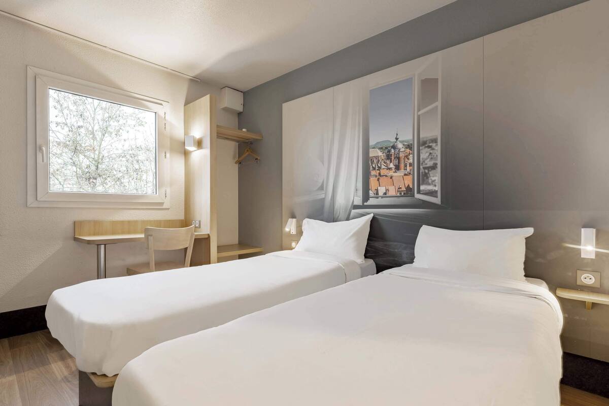 A hotel room with two single beds, a small desk, a chair, and a large window with a cityscape view.