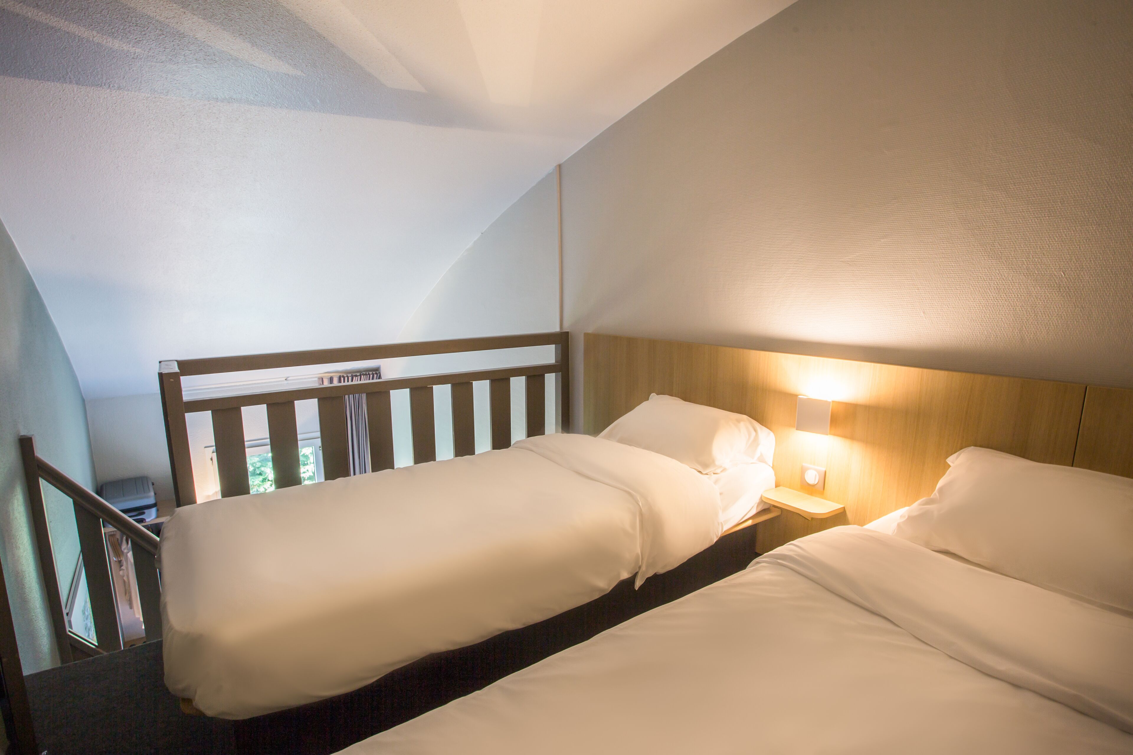 A hotel room with two single beds, a wooden headboard, a nightstand with a lamp, and a view of the outdoors through a window.