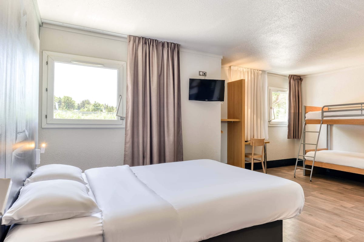 Une chambre d’hôtel avec un grand lit, un téléviseur fixé au mur, une fenêtre avec des rideaux et un lit superposé dans un coin.