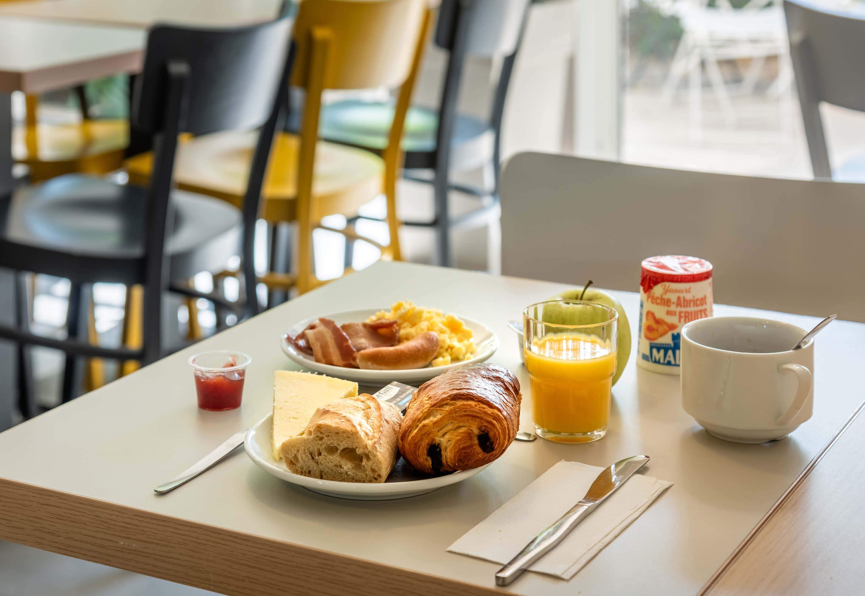 Une table de petit-déjeuner avec des croissants, des œufs, du bacon, un verre de jus d’orange, une tasse de café et un petit pot de confiture.