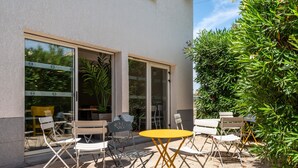 Un espacio al aire libre con una mesa amarilla y sillas blancas, rodeado de follaje y un edificio con ventanas amplias.