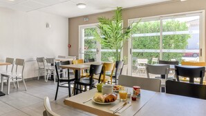 Comedor con mesas y sillas, una planta y una ventana que da a vistas de follaje y un coche estacionado afuera.