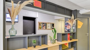 Un mostrador de recepción de diseño moderno, con un jarrón de vidrio con plumas de miscania, un cactus y una pequeña planta en maceta. Encima del mostrador hay una pizarra de menú enmarcada y una puerta con un letrero que indica un área privada local.