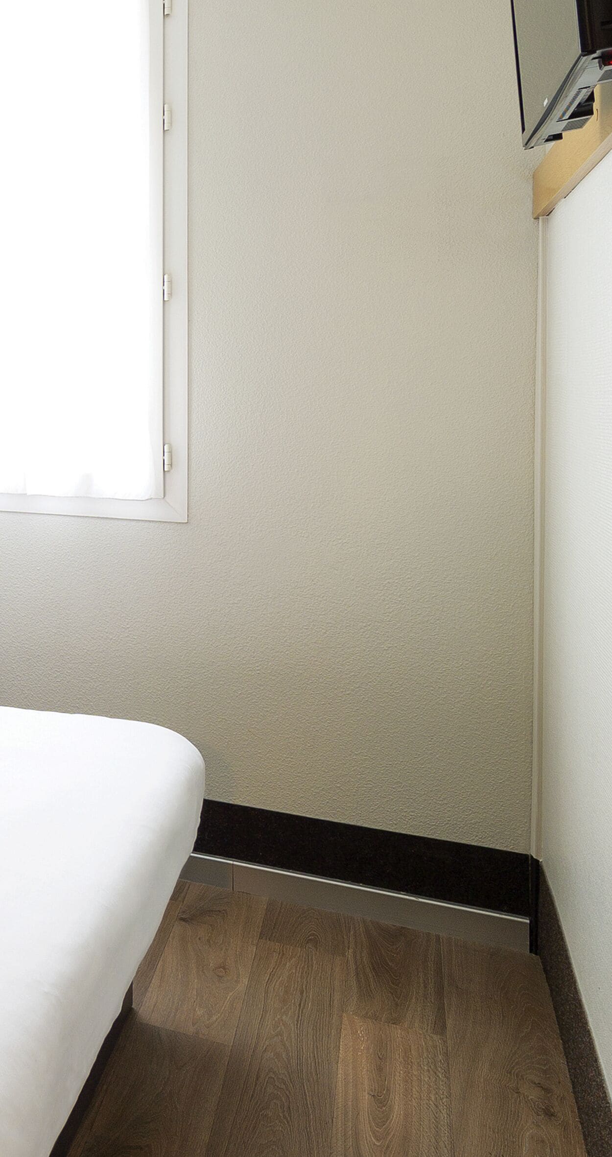 A corner of a room with a white bed, a wooden floor, and a white wall.