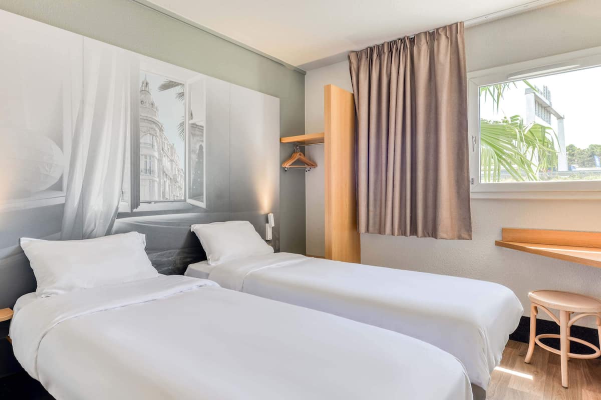 Une chambre d’hôtel avec deux lits, un bureau en bois, un porte-manteau et une fenêtre avec des rideaux.