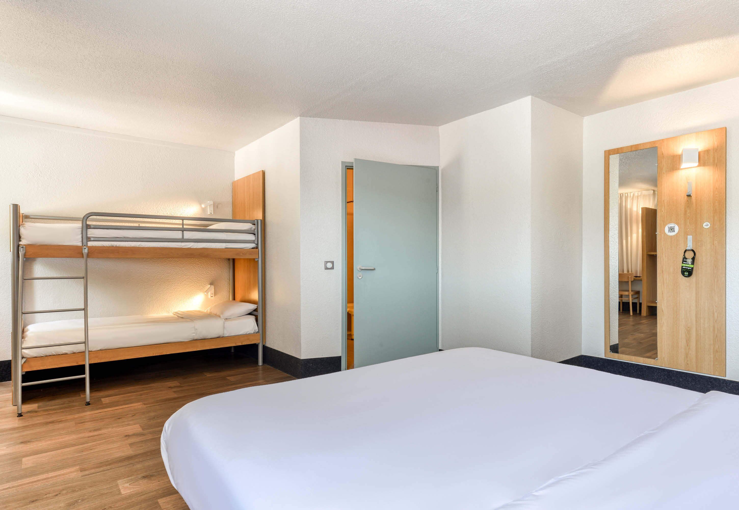 A hotel room with a bunk bed, a single bed, a wooden wardrobe, a door, and a mirror.