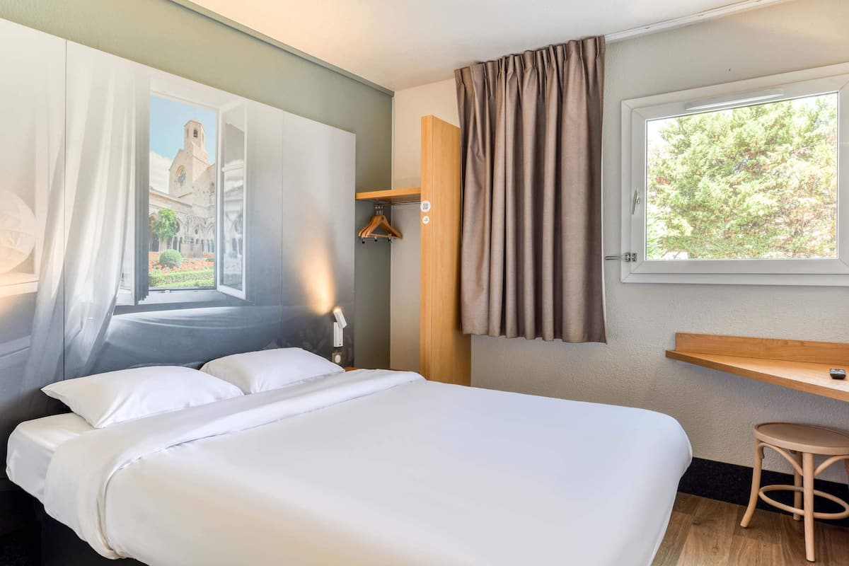 Une chambre d’hôtel avec un lit, un bureau, une chaise, une fenêtre avec des rideaux et un tableau au mur.