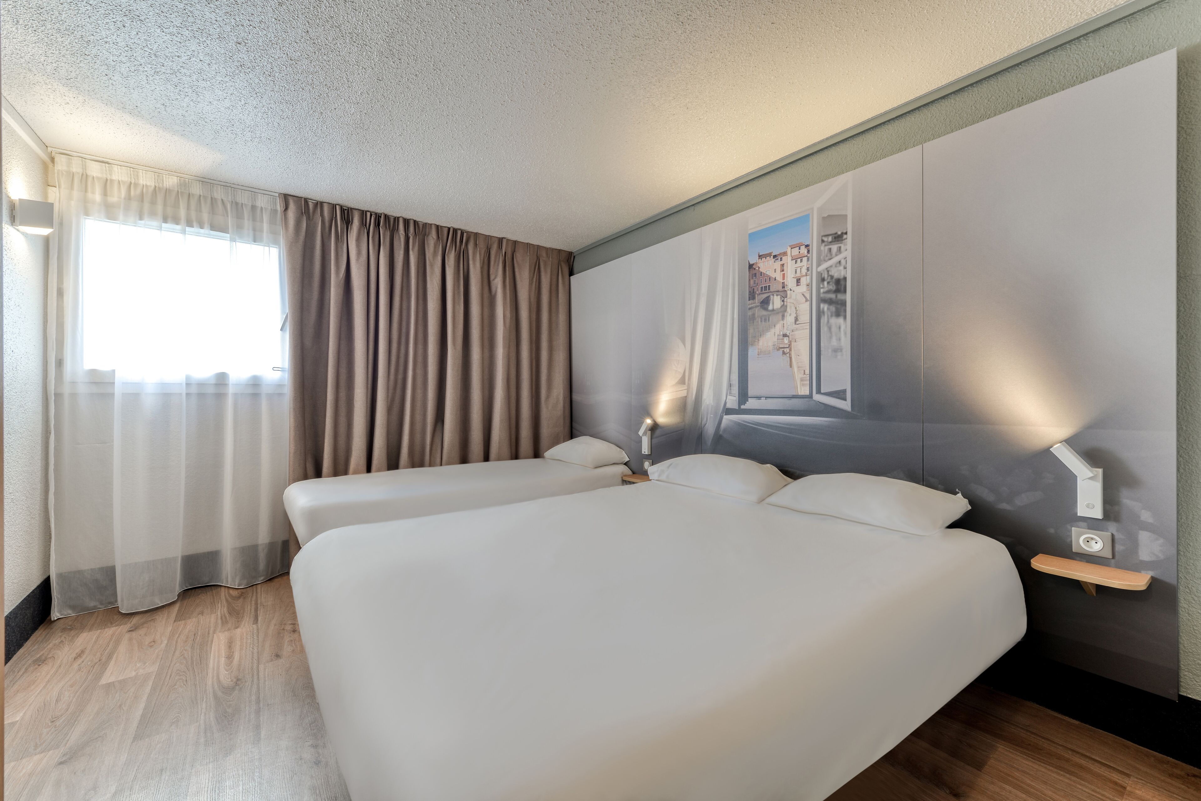 Habitación de hotel con dos camas, un ventanal con cortinas, una lámpara en la pared y suelo de madera.