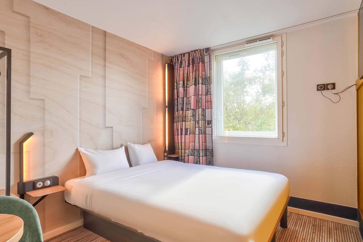 A neatly made bed with white linens, a bedside table with a lamp, and a window with patterned curtains.