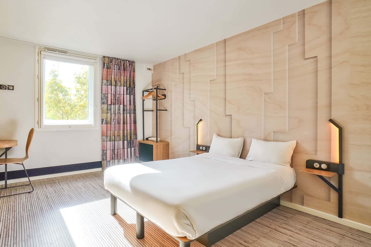 A modern hotel room with a large bed, a desk, and a chair. There is a window with curtains, a nightstand with a lamp, and a wall with a geometric pattern.