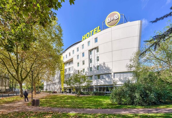 A modern hotel building with a prominent "HOTEL" sign, surrounded by trees and a well-maintained lawn. - B&B HOTEL Paris Malakoff Parc des Expositions (Malakoff)