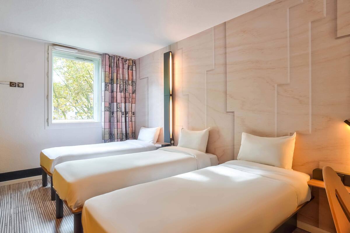 A hotel room with two beds, a window with curtains, a wall-mounted light, and a patterned curtain.