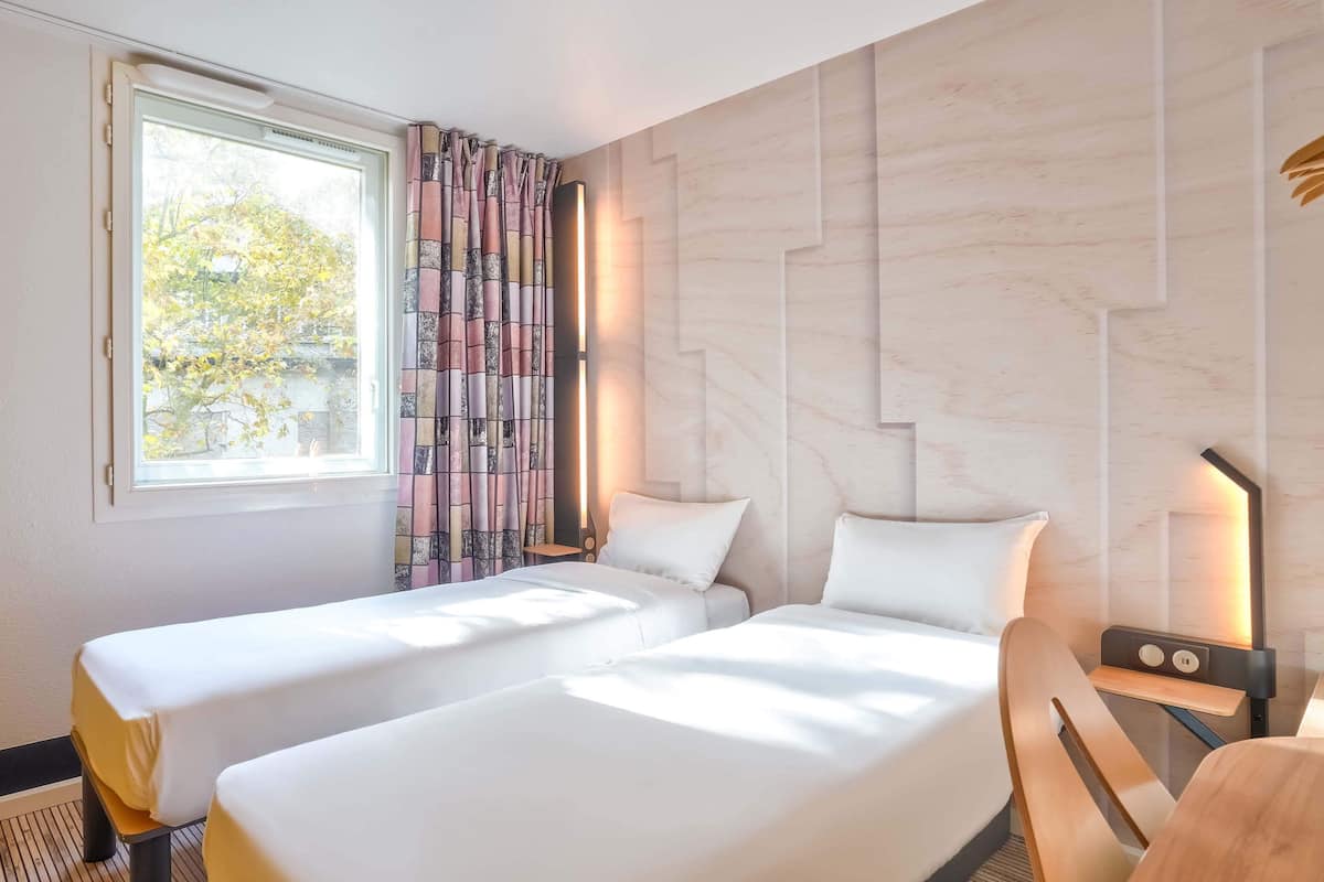 A hotel room with two beds, a window with curtains, a wall with a geometric design, and a bedside table with a lamp.