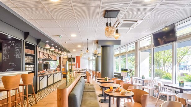 A modern cafe with a long counter, seating area, and large windows. - B&B HOTEL Paris Malakoff Parc des Expositions (Malakoff)