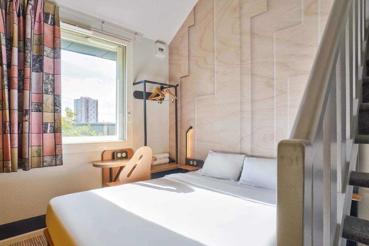 A hotel room with a bed, a desk, a chair, a window with curtains, and a view of buildings outside.