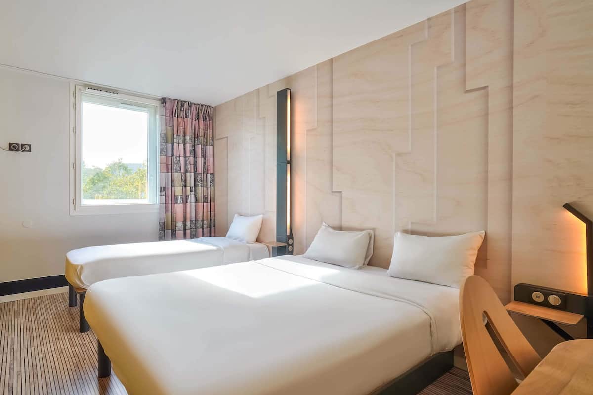 A hotel room with two beds, a window with curtains, a wall-mounted light, and a wooden chair.