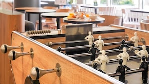 A foosball table in a brightly lit room with wooden furniture and large windows. - B&B HOTEL Paris Malakoff Parc des Expositions (Malakoff)