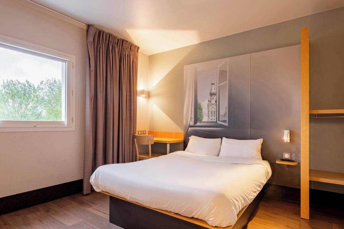 Una habitación de hotel moderna con una cama grande, un escritorio y una silla. Hay una ventana con cortinas, una lámpara de pared y un estante pequeño con una radio.