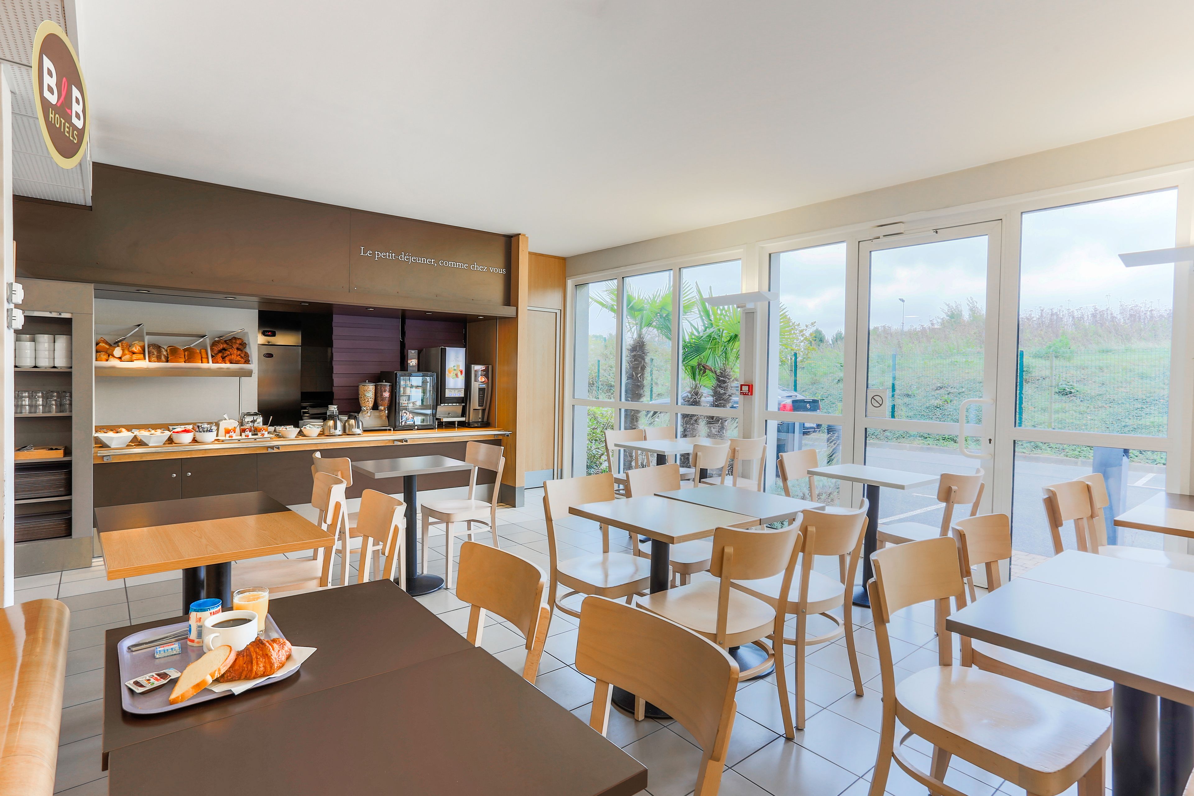 Un espace de restauration moderne dans un hôtel, avec des tables et des chaises, une cuisine en arrière-plan et de grandes fenêtres donnant sur l’extérieur.