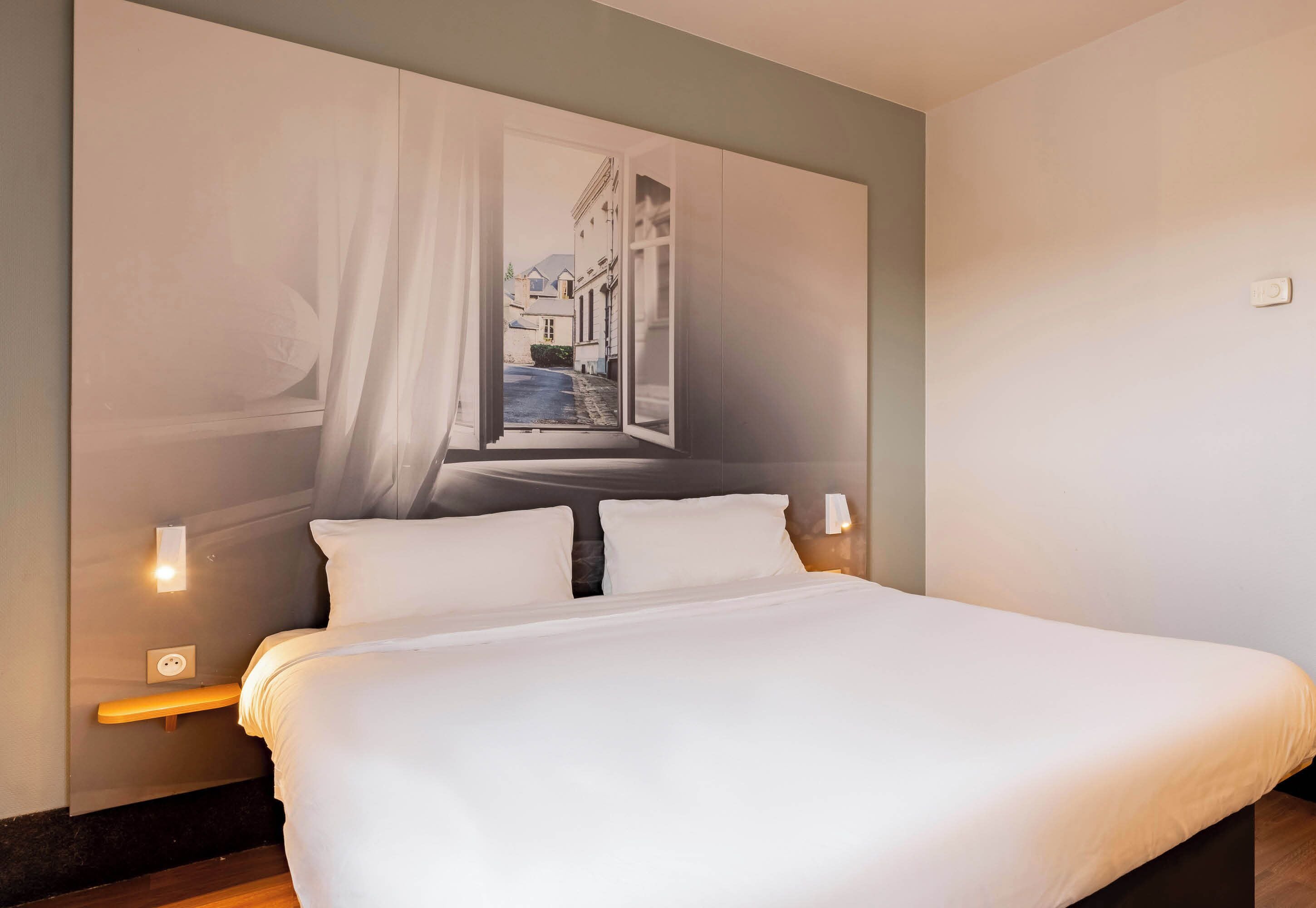 Una cama bien hecha con sábanas blancas, cabecera con un mural de paisaje urbano y lámparas de noche.