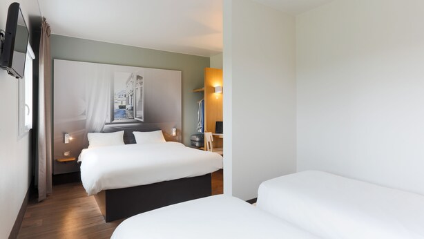 Habitación de hotel con dos camas, un televisor de pantalla plana montado en la pared, una mesita de noche con lámpara y un ventanal con cortinas.