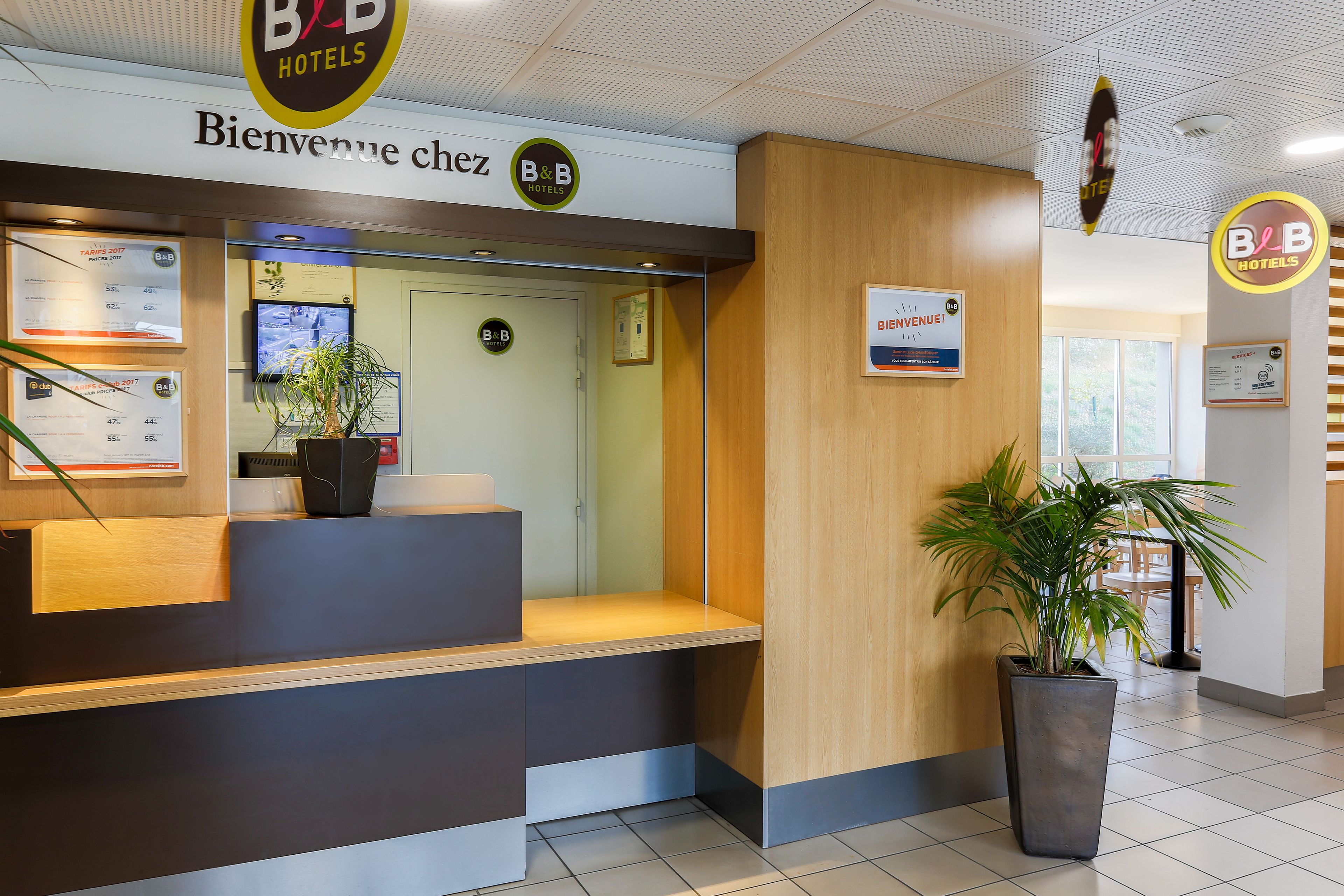 Ein Hotel-Empfangsbereich mit einer Rezeption, einer Pflanze und einem Schild, auf dem 'Bienvenue chez B&B HOTELS' steht.