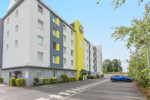 A multi-story hotel with a yellow and grey facade, a parking lot, and a blue car parked in front. - B&B Hotel Valenciennes Marly (Marly)