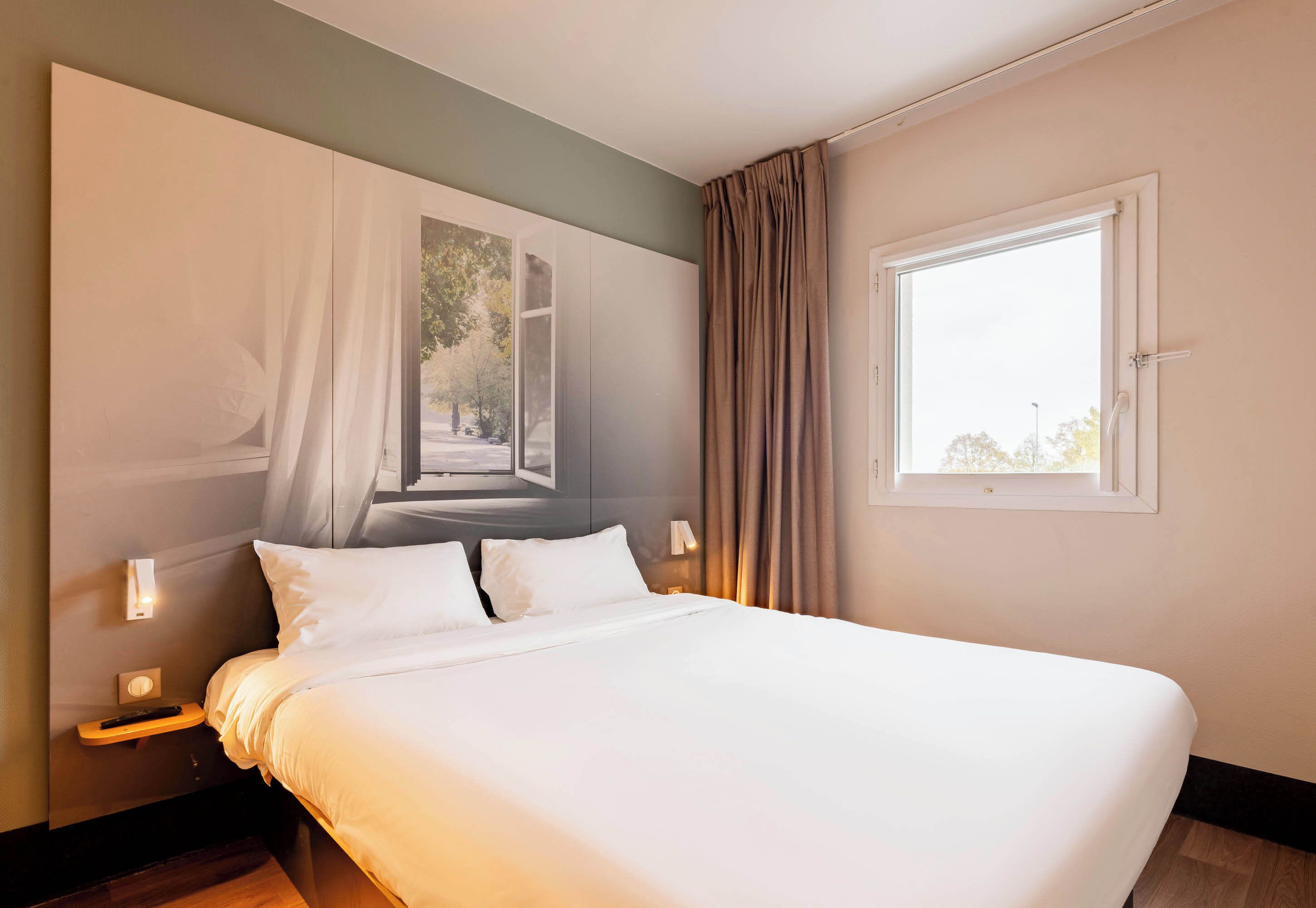 Een keurig opgemaakt bed in een hotelkamer met een groot raam, een nachtkastje met een lamp en een aan de muur hangend kunstwerk.