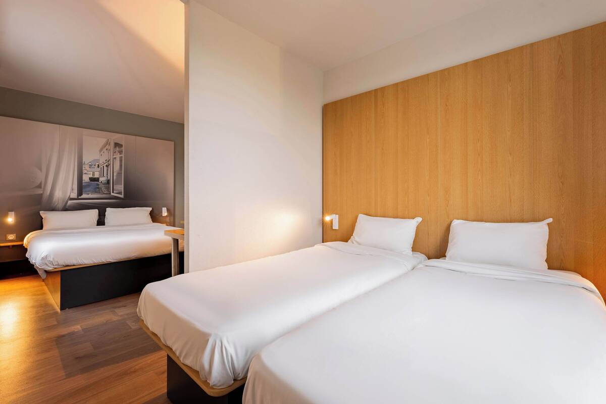 Habitación de hotel con dos camas, cabeceros de madera y un ventanal con vistas a edificios.