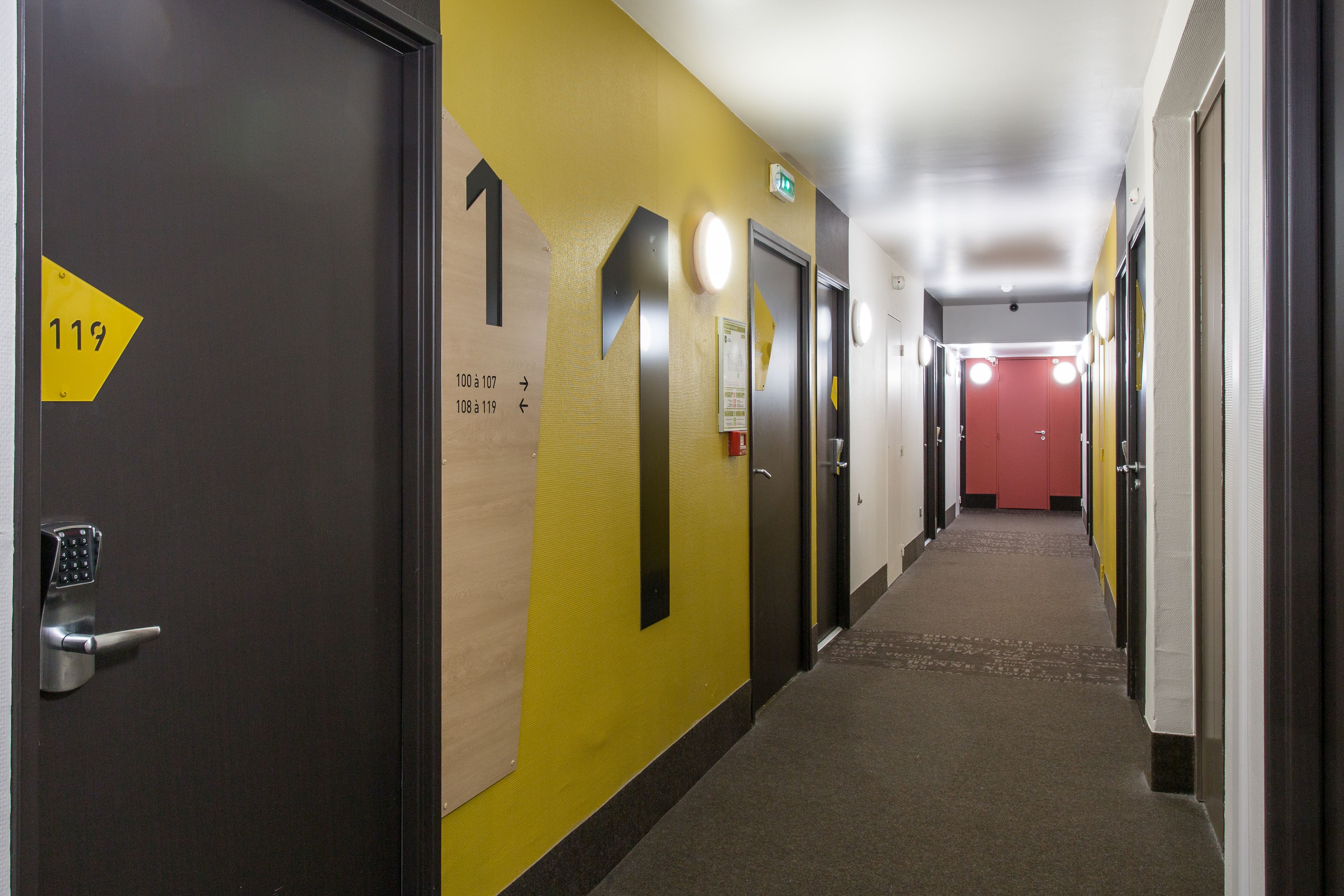 Ein Hotelkorridor mit Türen, die mit den Nummern 119 und 108 bis 119 gekennzeichnet sind, einer roten Tür am Ende und Pfeilen an den Wänden.