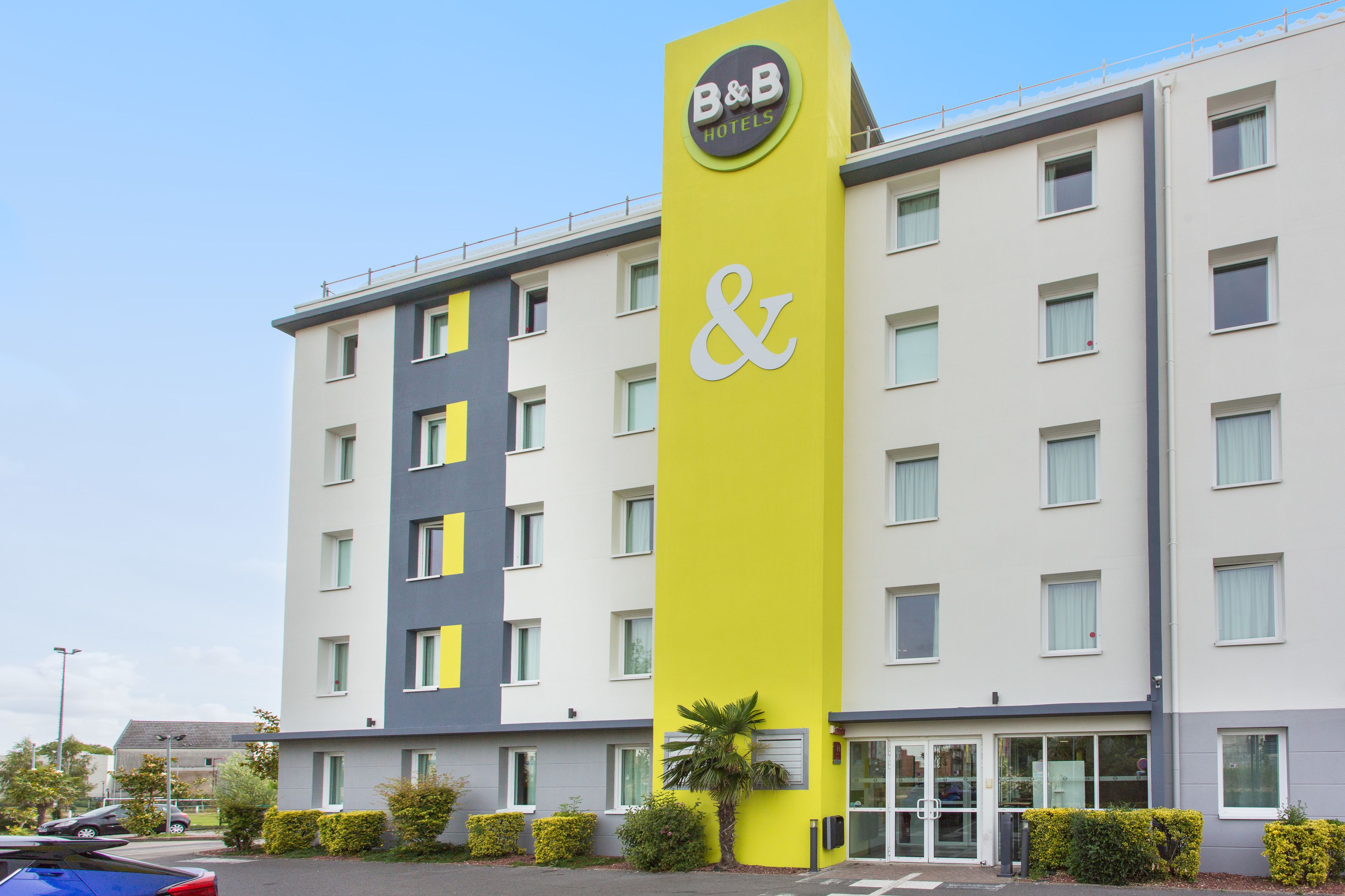 A B&B hotel with a yellow and grey facade, multiple windows, and a palm tree in the front.