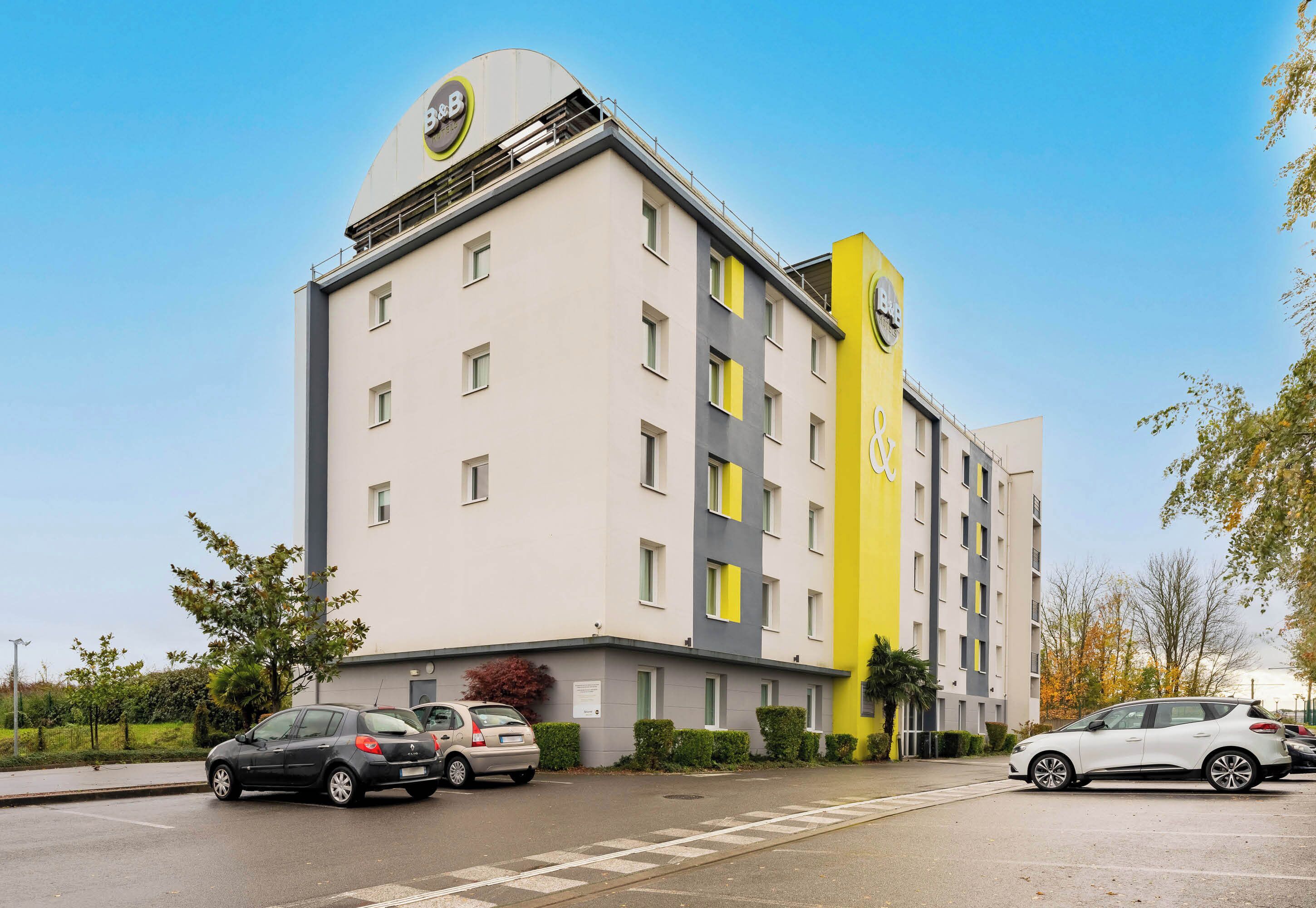 A multi-story hotel with a modern design, featuring a prominent yellow and gray facade, multiple windows, and a parking area with several cars.
