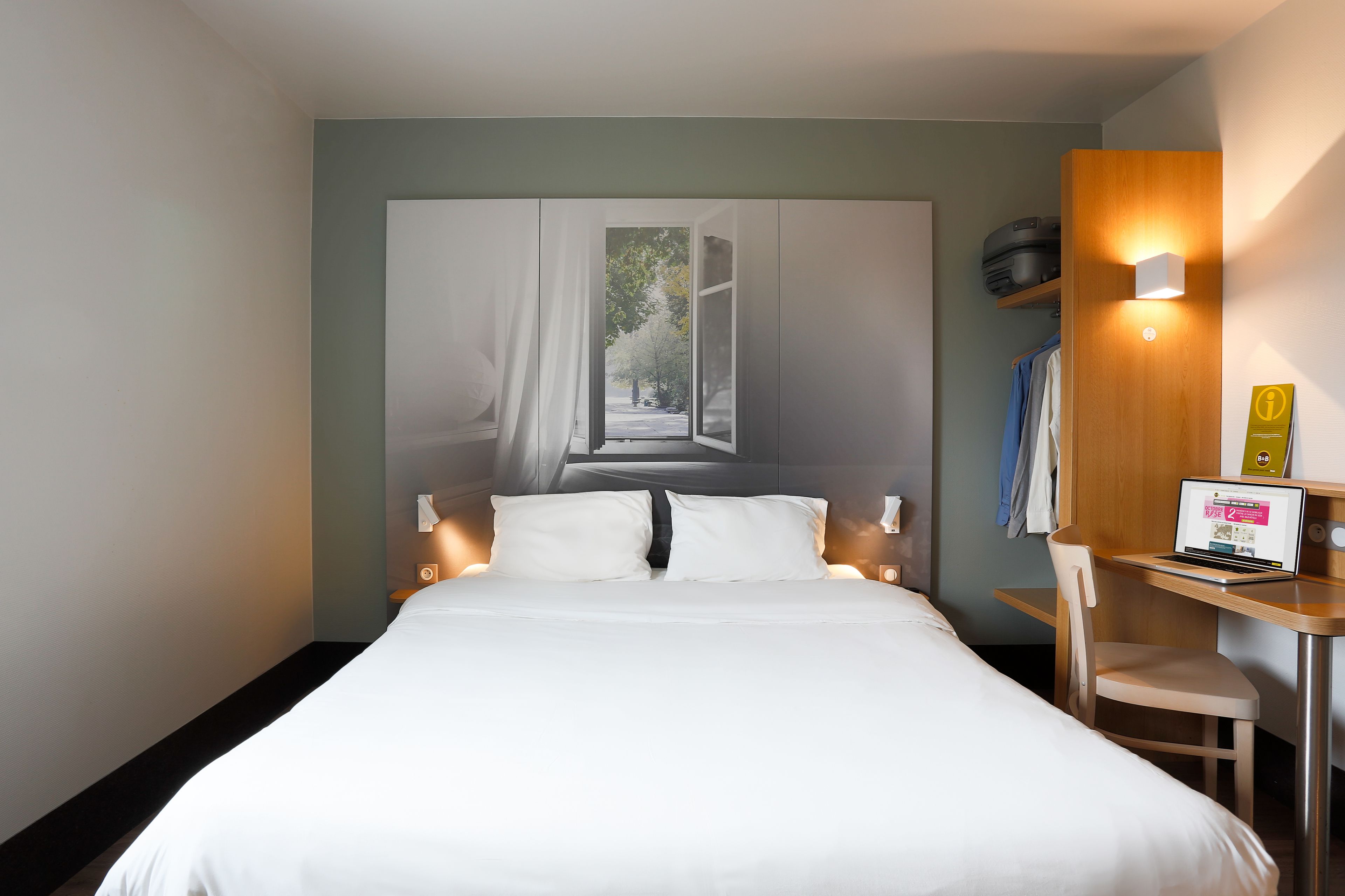 A hotel room with a bed, a desk with a laptop, a chair, and a large window with curtains.