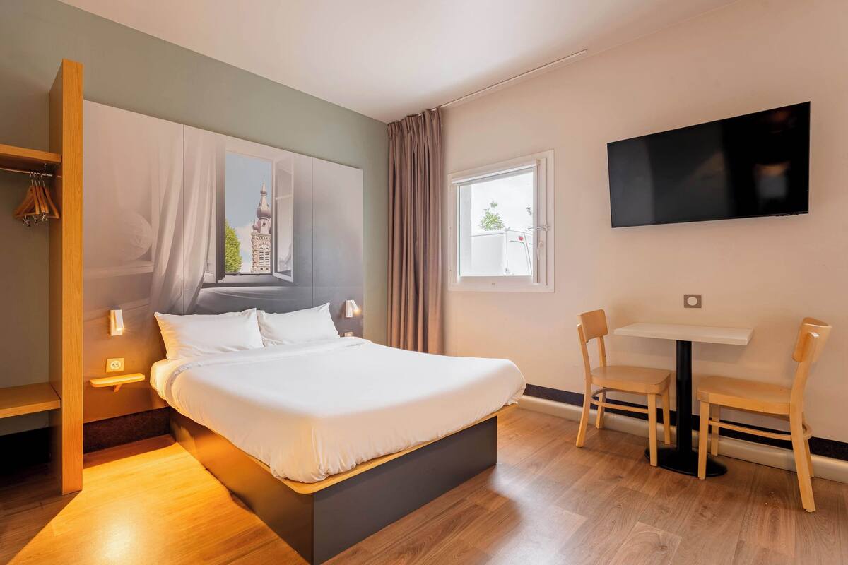 Habitación de hotel con cama, escritorio, sillas, televisor y ventana con vistas a un edificio.