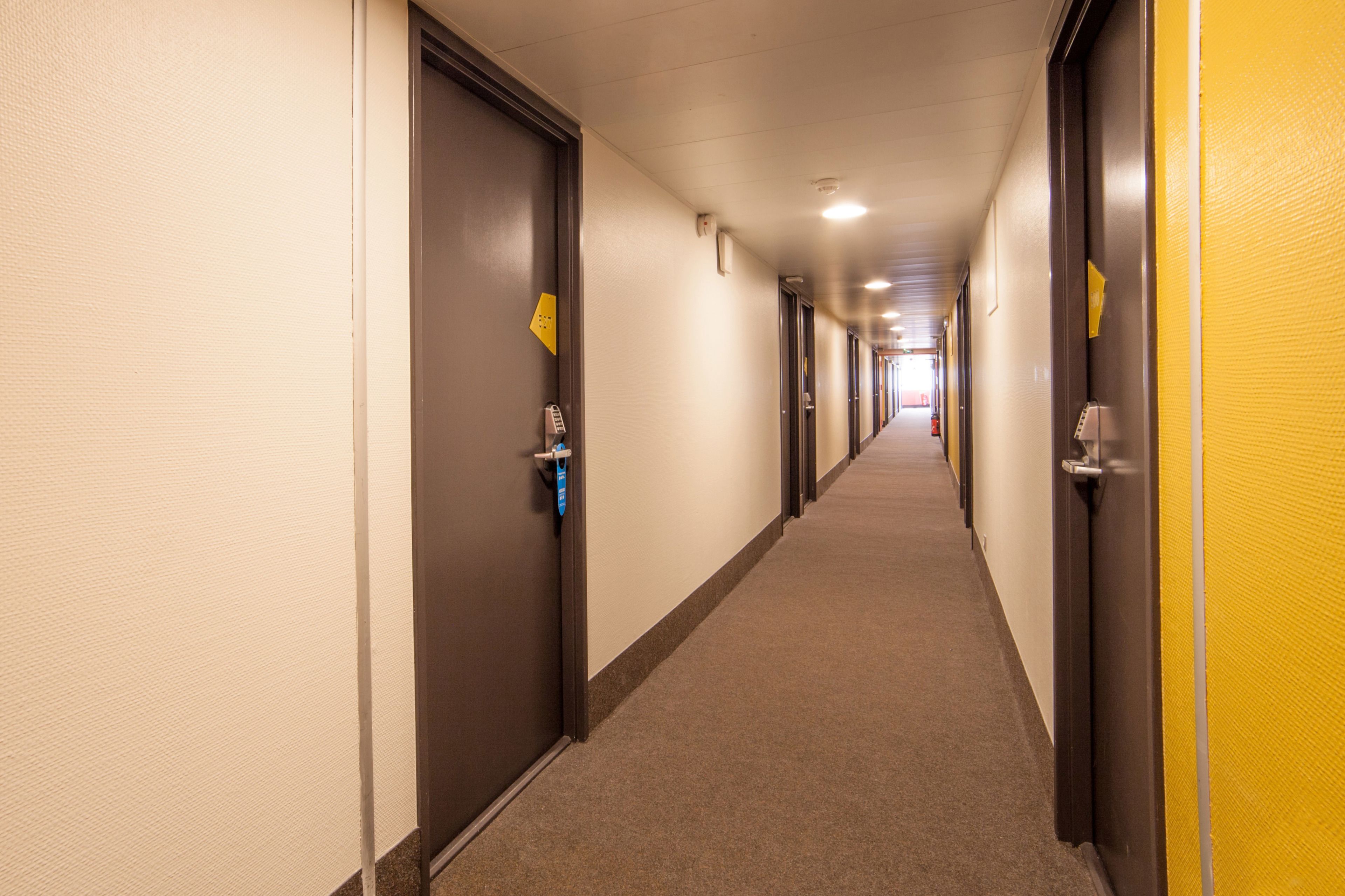 Un corridoio d'albergo con porte su entrambi i lati, un pavimento rivestito di moquette e una parete gialla.