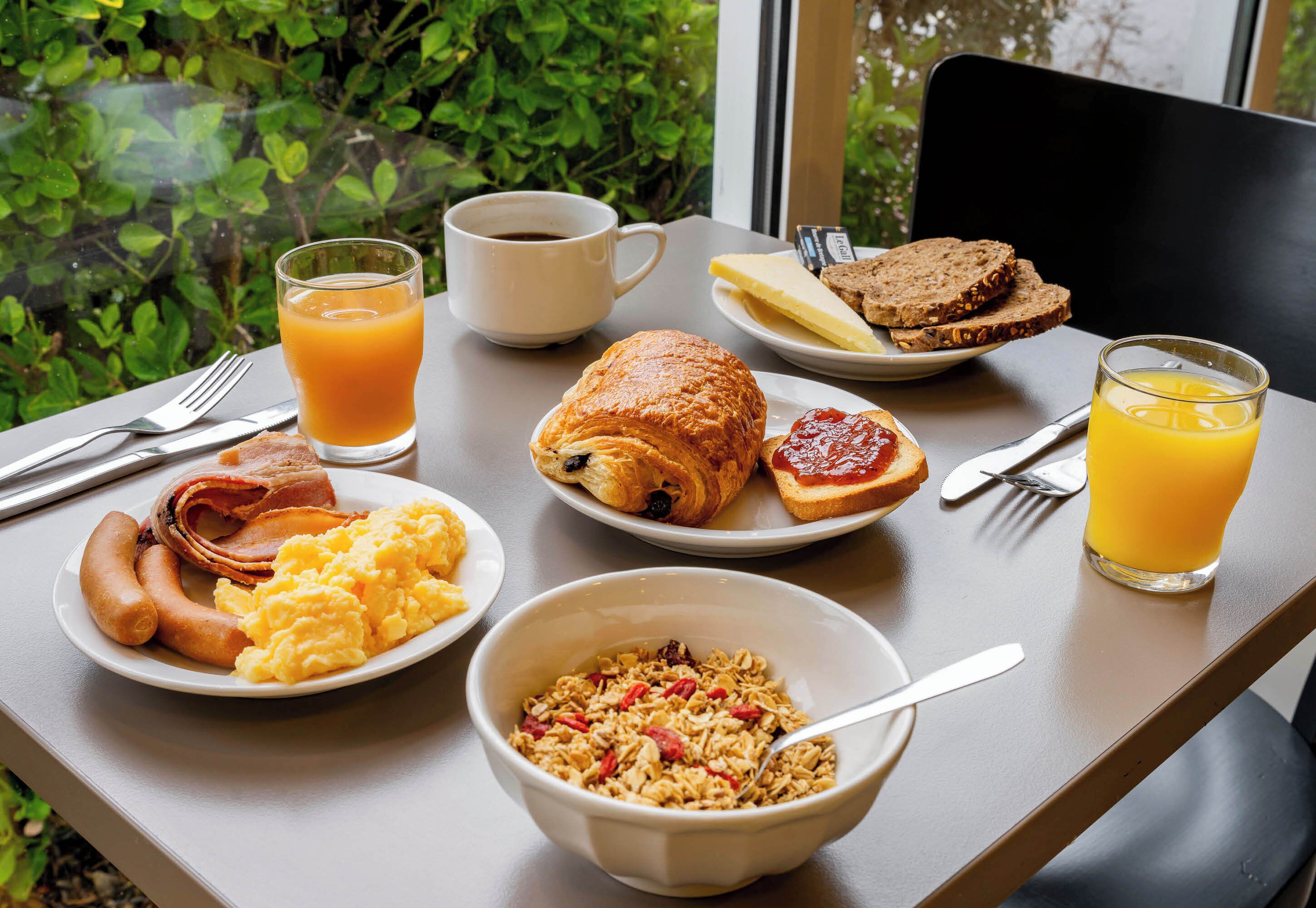 Una mesa de desayuno con platos de huevos, salchichas, croissants y tostadas, junto con un tazón de cereales y vasos de zumo de naranja.