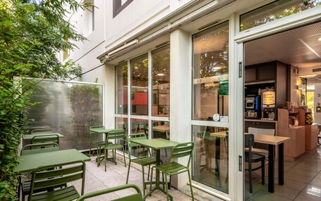 An outdoor seating area with green tables and chairs, a glass door entrance, and a view of the interior through the glass.