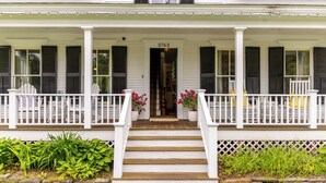 A white building with black shutters, a number '5763' above the entrance, and a porch with white railings and potted plants.