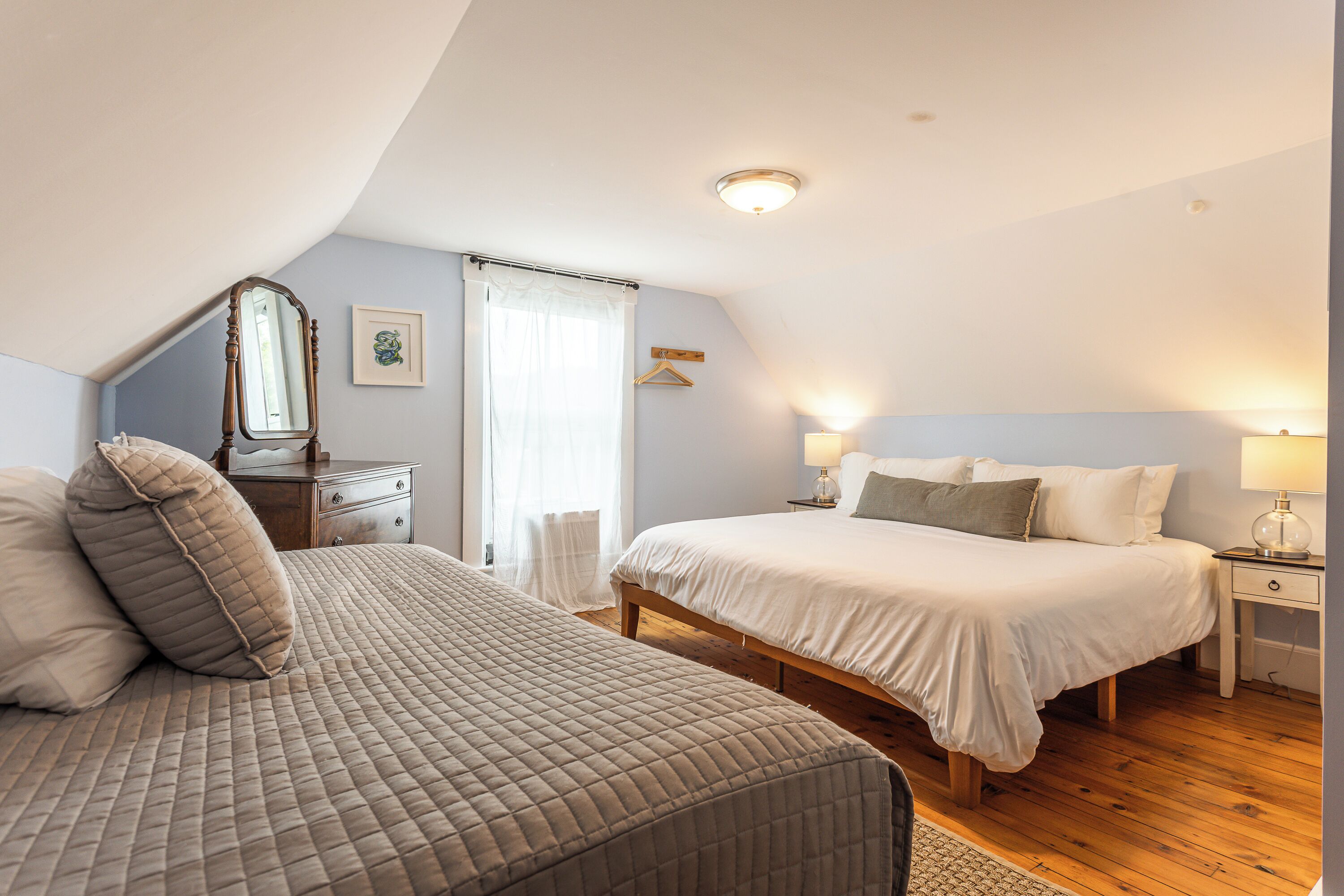 A bedroom with two beds, a dresser, a mirror, a bedside table with a lamp, and a window with curtains.