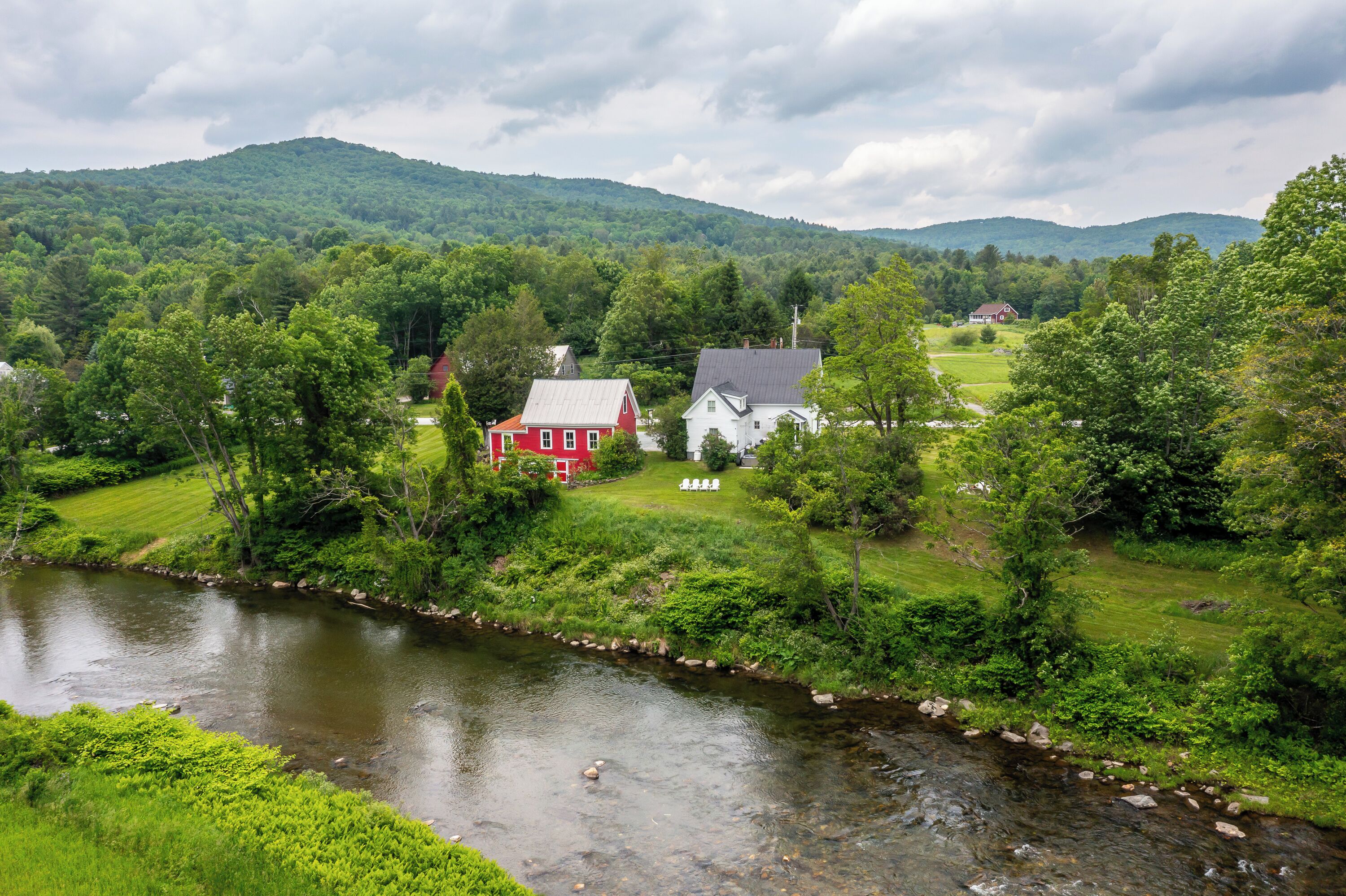 A river flowing through a lush green landscape with two houses, one red and one white, surrounded by trees and hills.