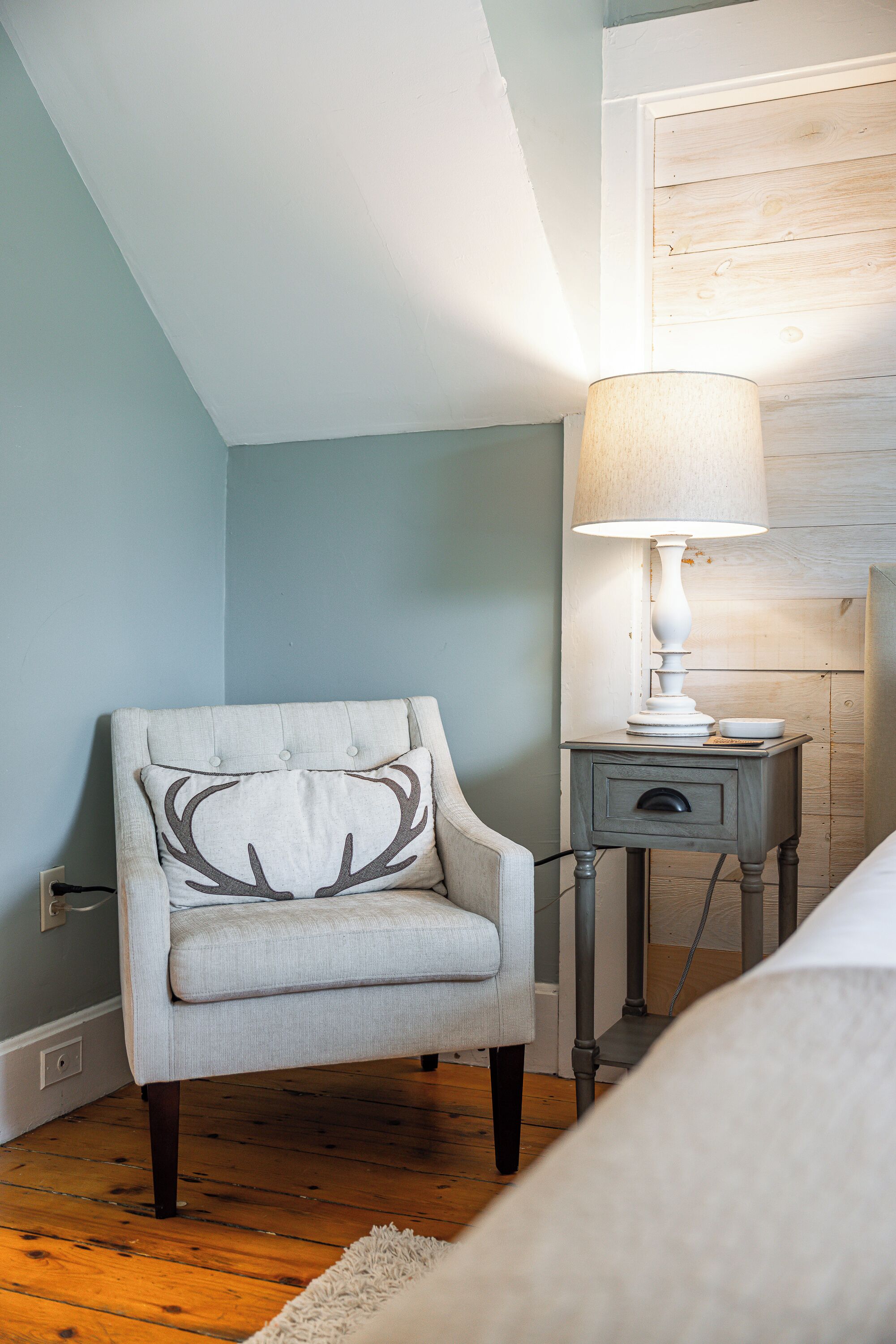 A bedroom with a light blue wall, a wooden floor, a white armchair with a deer pillow, a bedside table with a lamp, and a bed with a white blanket.