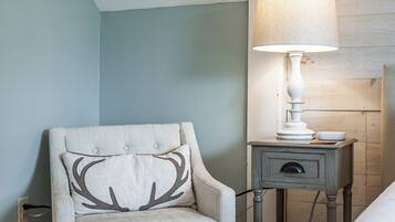 A bedroom with a light blue wall, a wooden floor, a white armchair with a deer pillow, a bedside table with a lamp, and a bed with a white blanket.