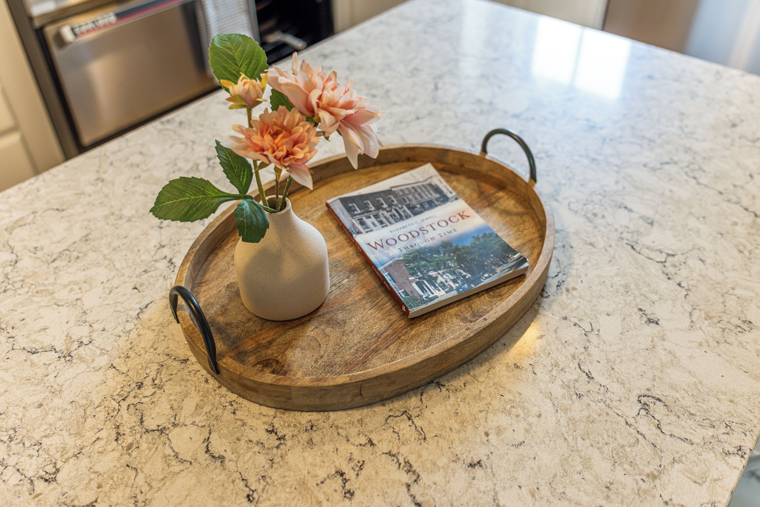 A wooden tray with a vase of flowers and a book on a marble countertop.
