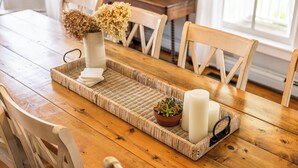 A wooden dining table with a woven tray, a vase with dried flowers, a small potted plant, and two candles.