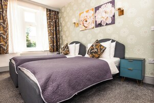 A double bed with a purple bedspread, a nightstand with a teal drawer, and floral wall art.