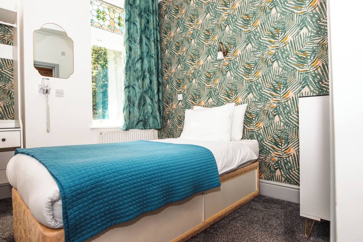 A bedroom with a bed, a mirror, a window with curtains, a radiator, and a wall with a leaf pattern.