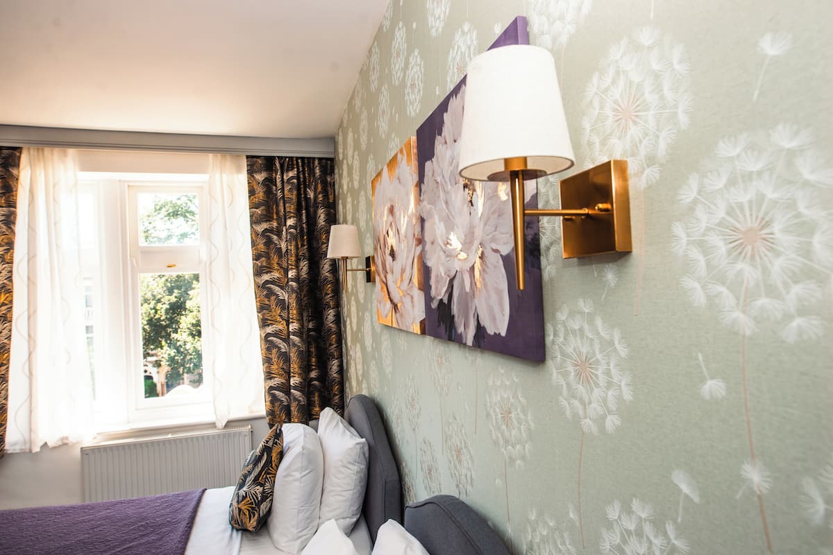 A hotel room with a bed, a window with curtains, a wall-mounted lamp, and a floral artwork.