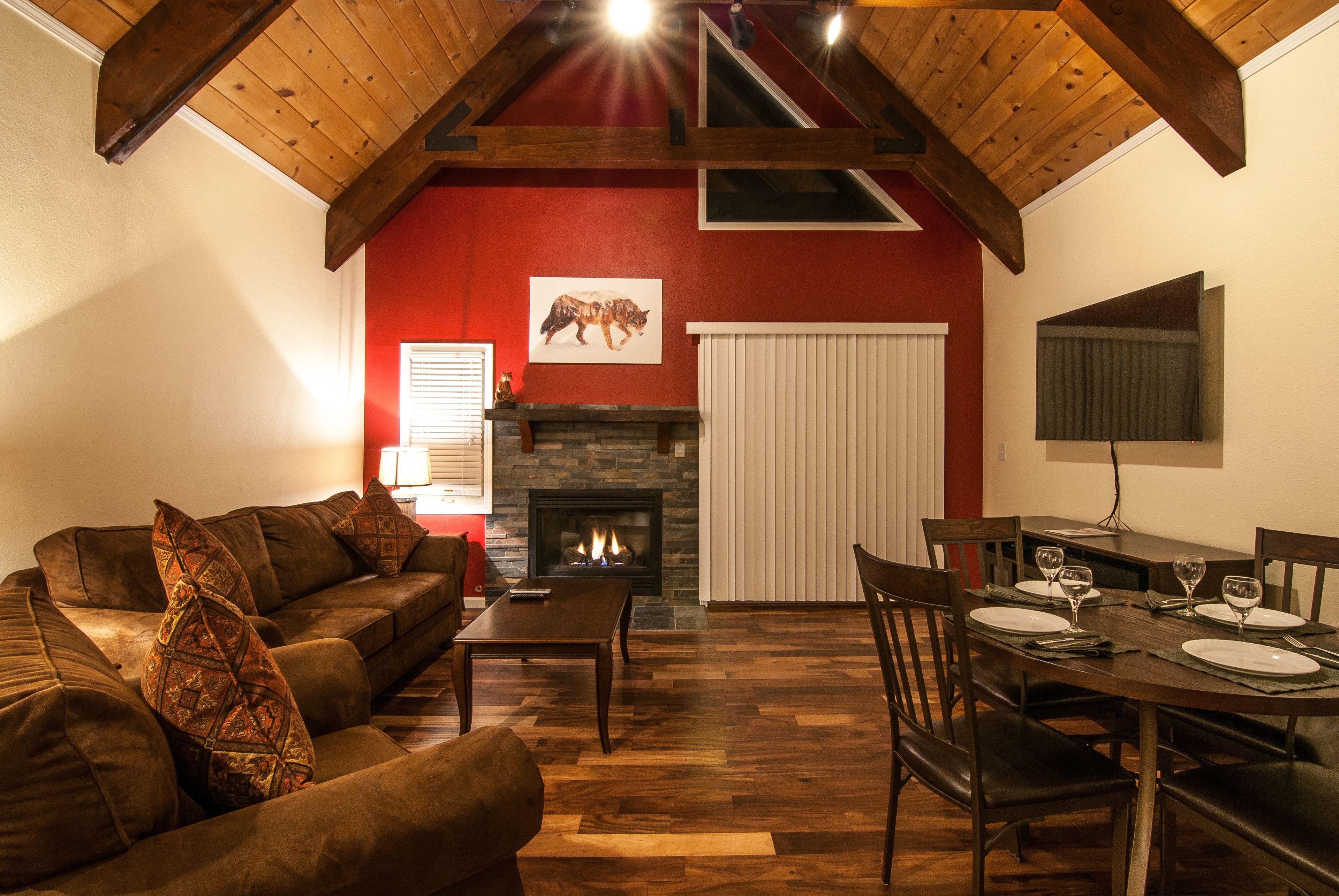 A living room with a fireplace, wooden ceiling beams, a dining area with a table set for four, and a red accent wall.