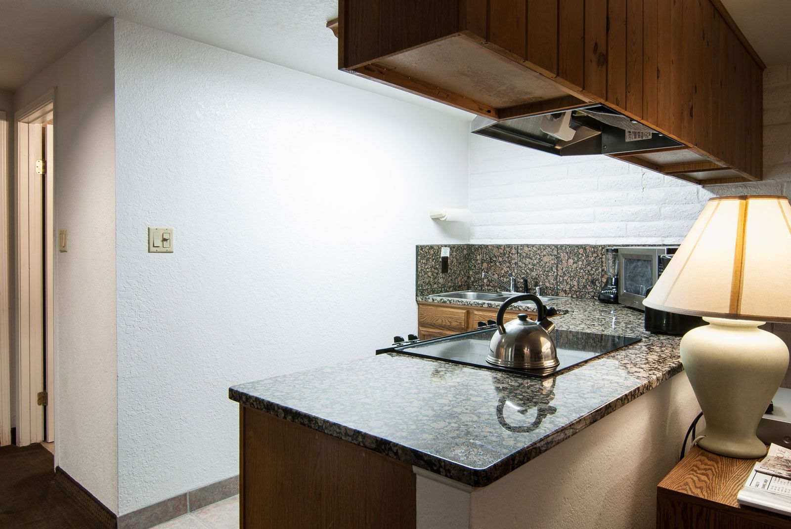 A compact kitchen with granite countertops, a kettle, a sink, and wooden cabinets.