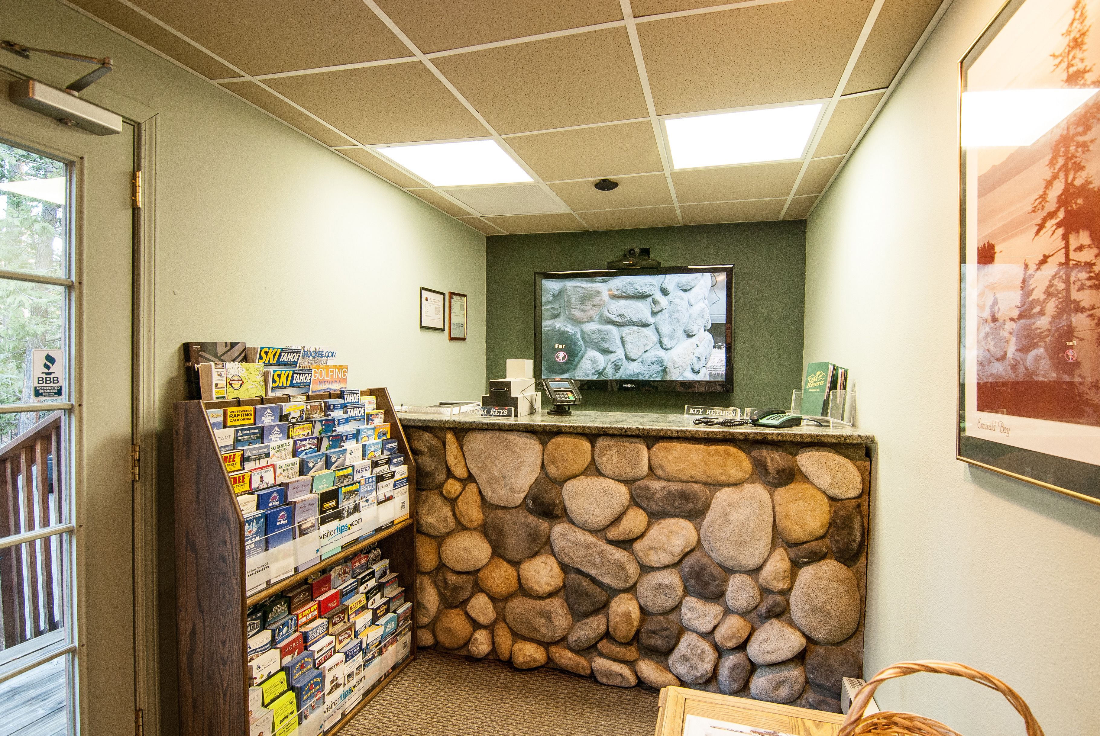 Zona de recepción con pared de piedra, mostrador con una computadora y exhibición de revistas.